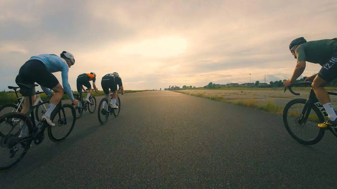 A group of cyclists in athletic gear ride their road bikes along a paved path under a soft, golden-hued sky.